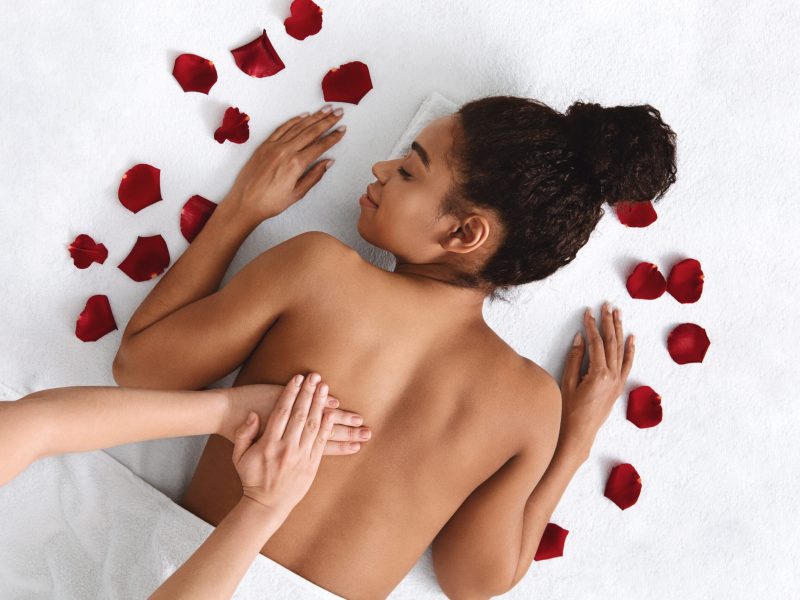 African girl getting healing back massage at spa decorated with rose petals, top view