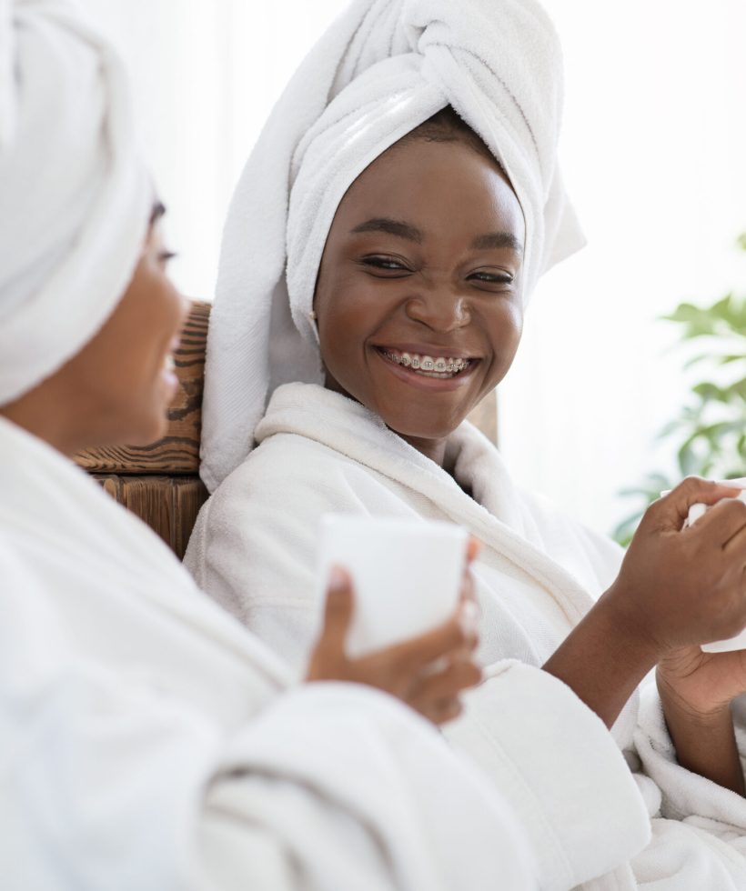 Closeup of two african american young women enjoying spa day at home, copy space. Two cheerful black ladies having conversation and drinking healthy tea while sitting on bed at home, side view